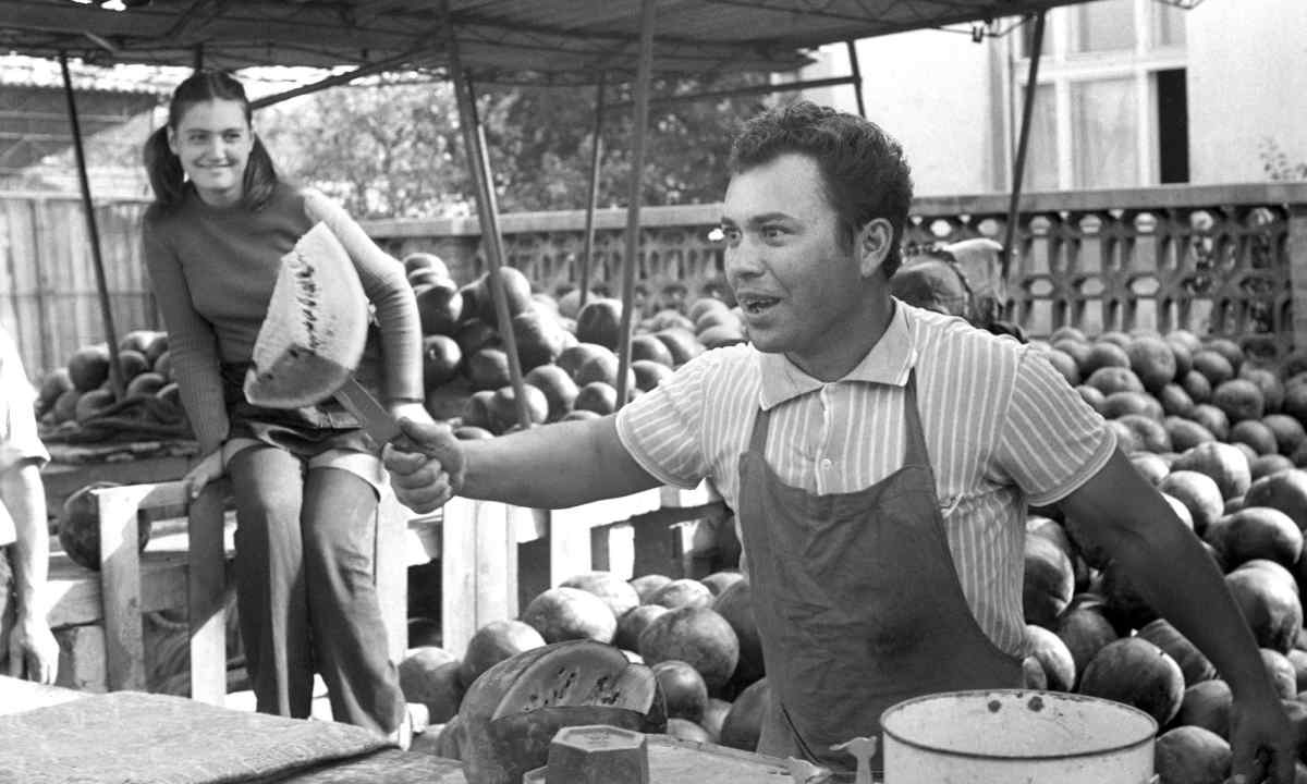 Guy holding a piece of a watermelon with his knife. In the background a girl is watching